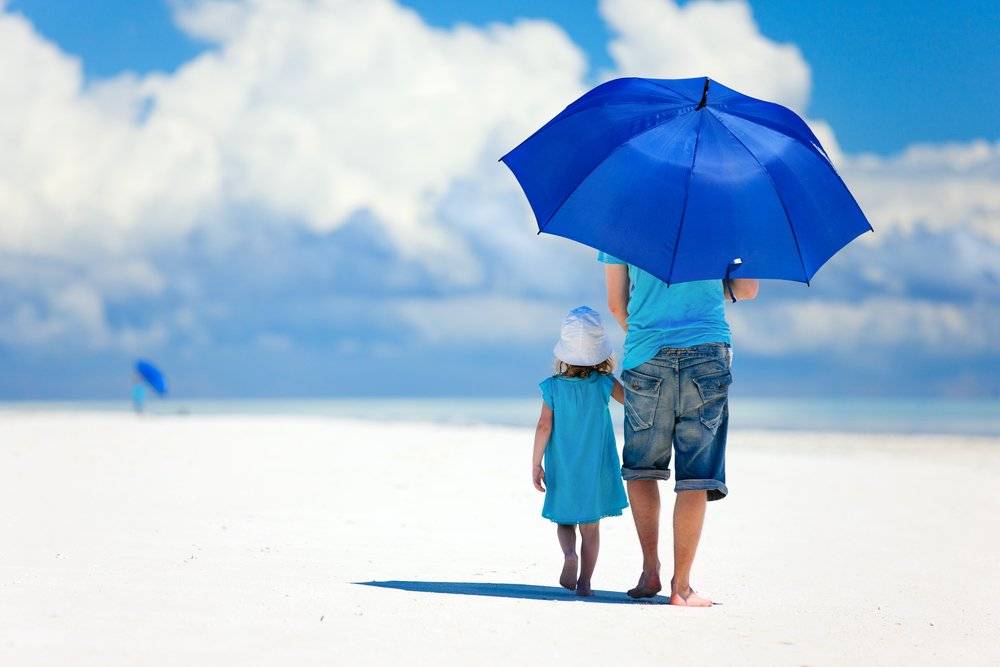 a parent and their child walking on a white sandy beach with a blue umbrella